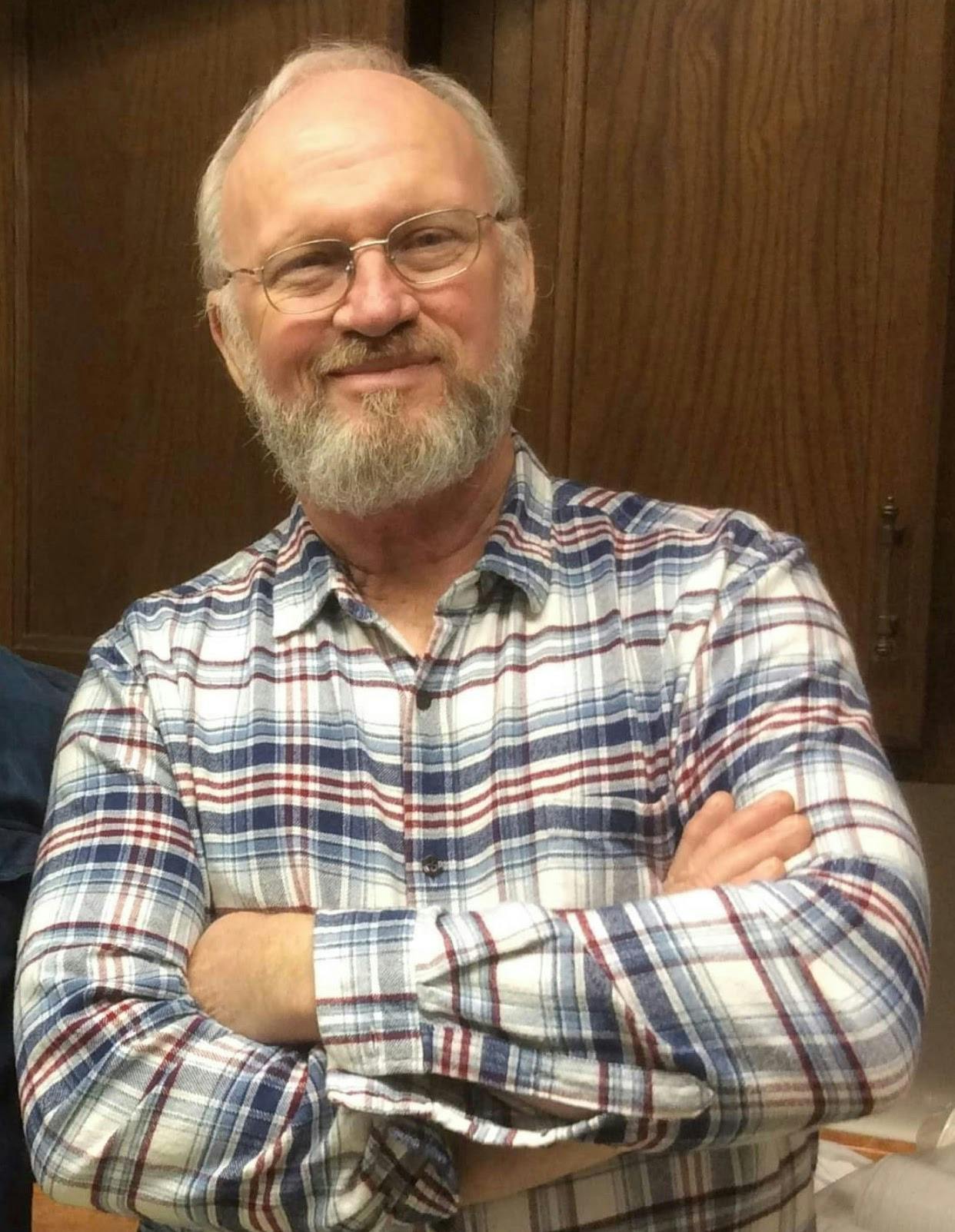 A photo of Joe Pursell, Board member and designer, standing with crossed arms and a plaid shirt.