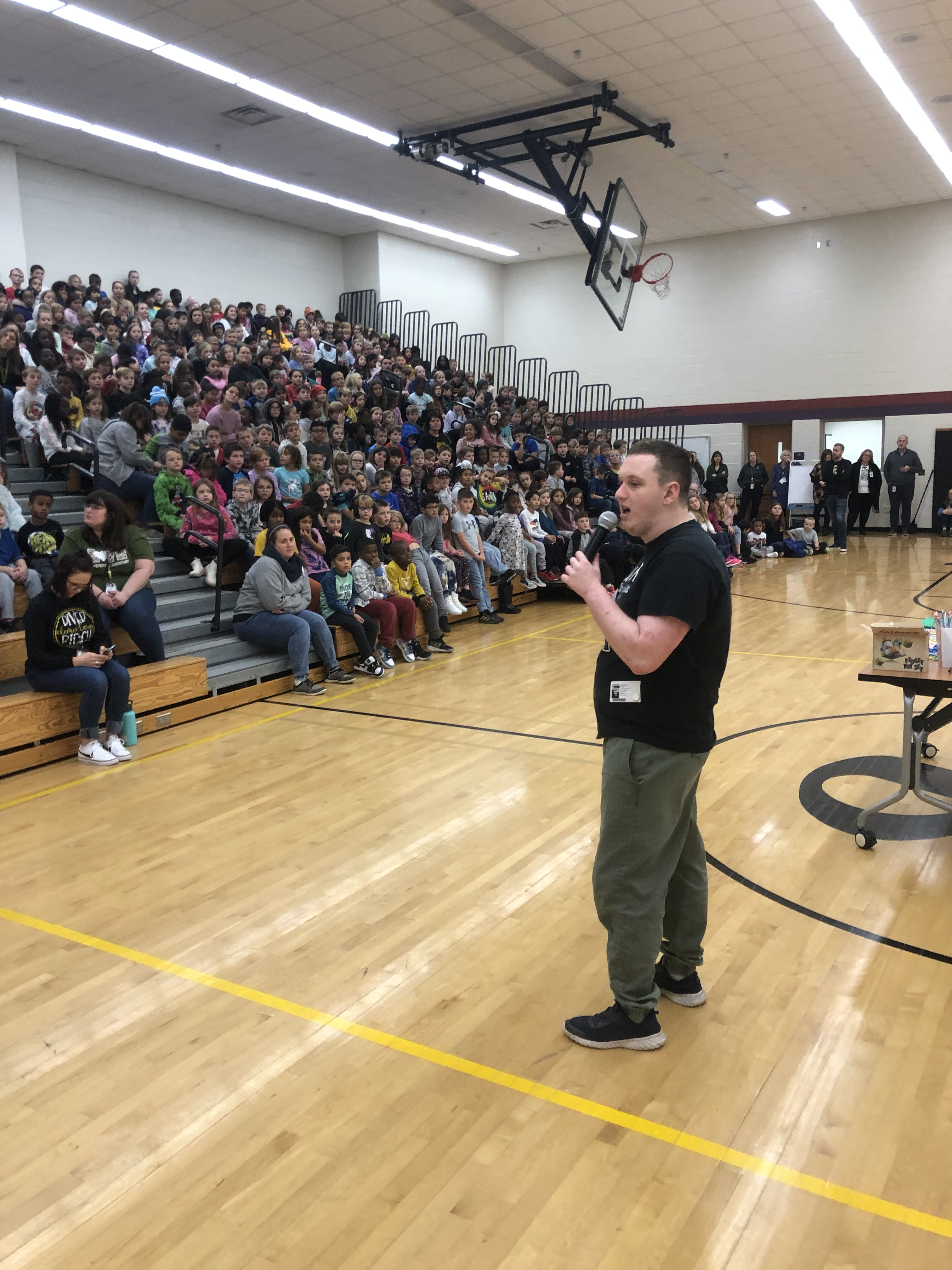Sam stands in the middle of a full gymnasium, speaking into a microphone to a crowd of teenagers.